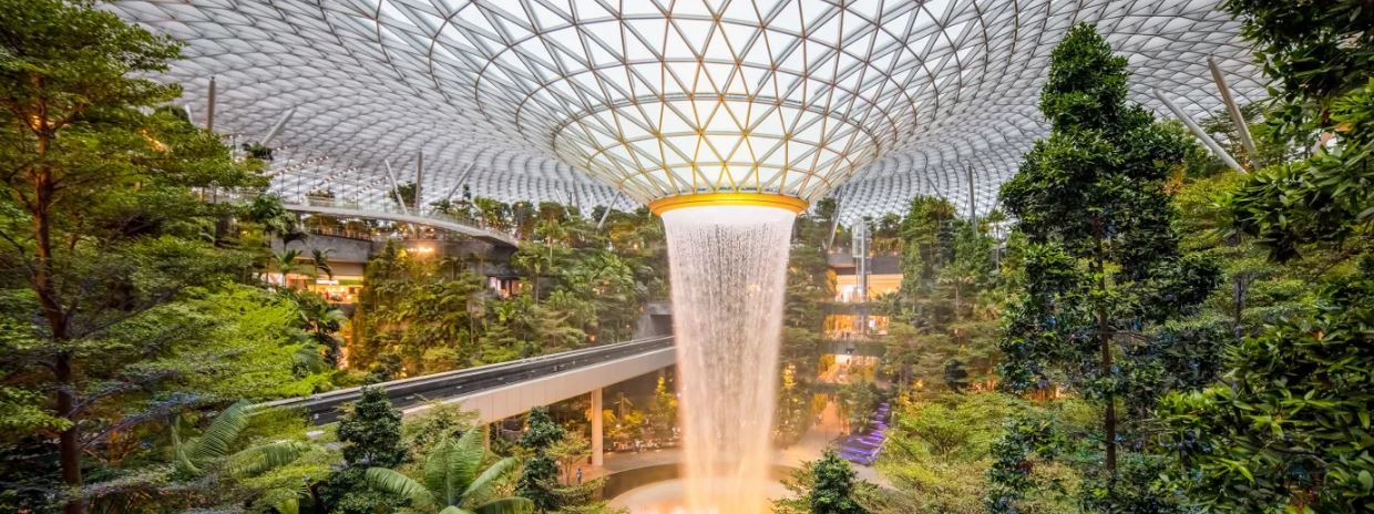 This meme is a split-screen image of the Jewel Changi Airport in Singapore. The top image shows a stunning, sprawling interior with lush greenery and a massive waterfall. The bottom image shows the same space, but with a giant, giant crystal – the world's largest – that has been installed at the airport. The text in the meme reads: “Jewel Changi Airport – where you can find both a giant crystal and a giant waterfall.”